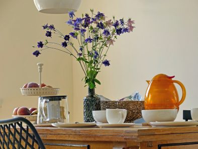 Auf dem Küchentisch der Ferienwohnung steht Geschirr, ein Blumenstraußen und eine Etagere mit Obst.