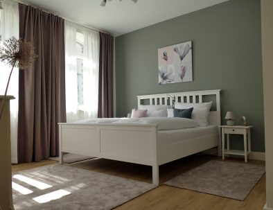 Das große Schlafzimmer in der Ferienwohnung Rhein-Lahn-Glück mit Doppelbett, Teppichen, Gardinen, Bild, Nachttisch und Nachttischlämpchen.