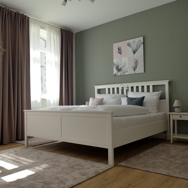 Das große Schlafzimmer in der Ferienwohnung Rhein-Lahn-Glück mit Doppelbett, Teppichen, Gardinen, Bild, Nachttisch und Nachttischlämpchen.