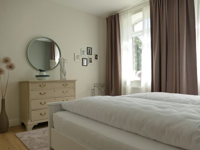 Das Schlafzimmer der Ferienwohnung Rhein-Lahn-Glück mit anitker Kommode, rundem Spiegel, kleiner Loreley-Bildergalerie und Doppelbett.