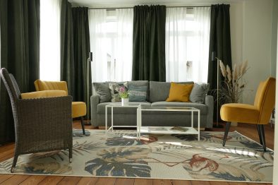 Das große Wohnzimmer der Ferienwohnung Rhein-Lahn-Glück mit grauem Sofa, Flamingo-Teppich und weißem Glastisch.