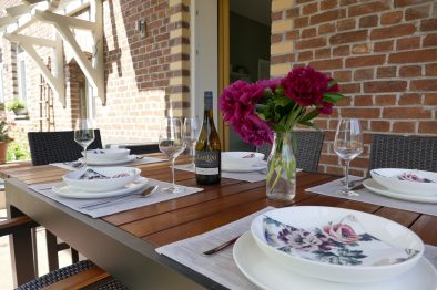 Der Terrassentisch der Mittelrhein-Ferienwohnung, gedeckt mit weißem Porzellan, Gläsern, einer Flasche Wein vom Weingut Laquai und einer Blumenvase.
