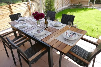 Terrasse der Mittelrhein-Ferienwohnung mit ausziehbarem Holztisch und sechs Gartenstühlen, gedeckt für sechs Personen. Auf dem Tisch stehen Blumen und eine Flasche Wein des Weingutes Laquai.