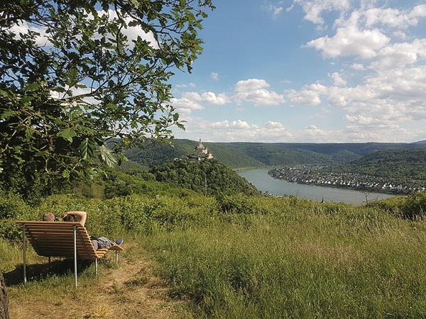 Auf einer geschwungenen Aussichtsbank sitzt ein Rheinsteig-Wanderer und blickt auf den Rhein und die Marksburg.