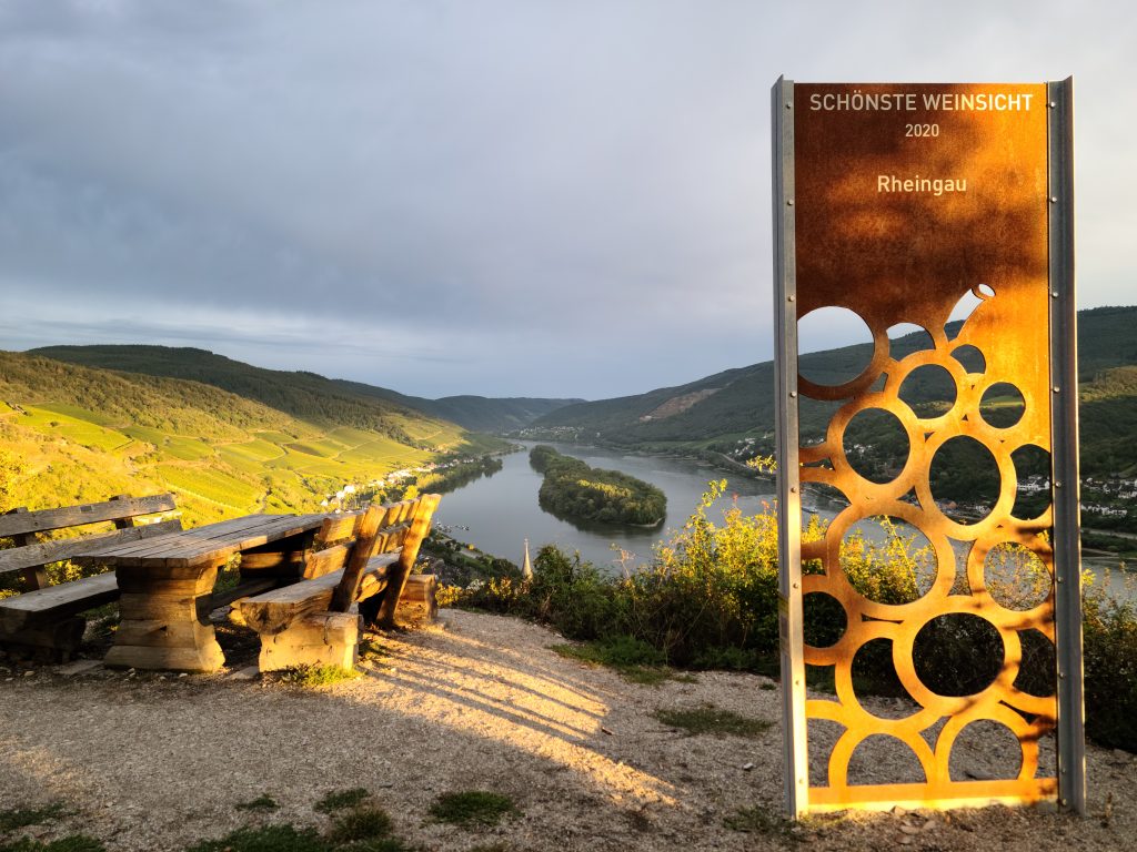 Eine große Metalltafel mit einer stilisierten Traube steht neben einer Picknickbank, im Hintergrund ist das Rheintal bei Lorch zu sehen.